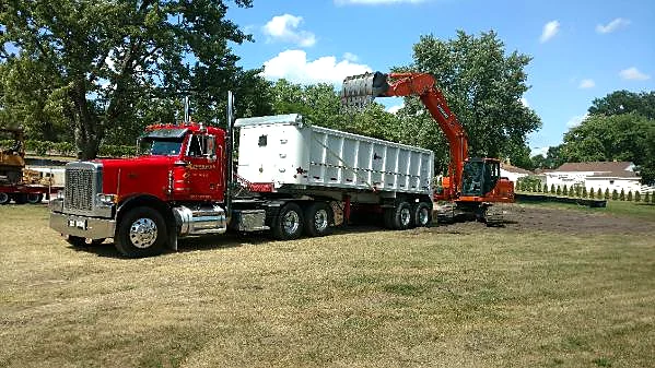 An excavator loading a red Krooswyk dump trailer on a jobsite